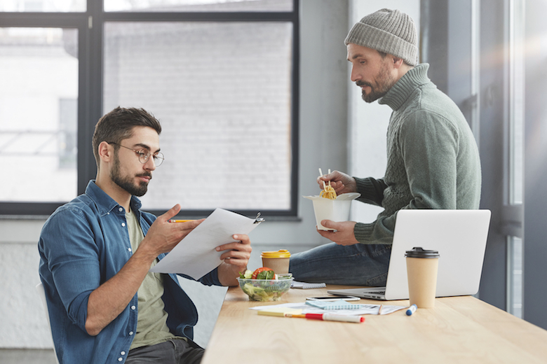 Dos hombres barbudos cooperan para hacer un proyecto común, almorzar, comer alimentos saludables rodeados de papeles y las tecnologías modernas hablan entre sí.  Concepto de cooperación y trabajo en equipo empresarial.